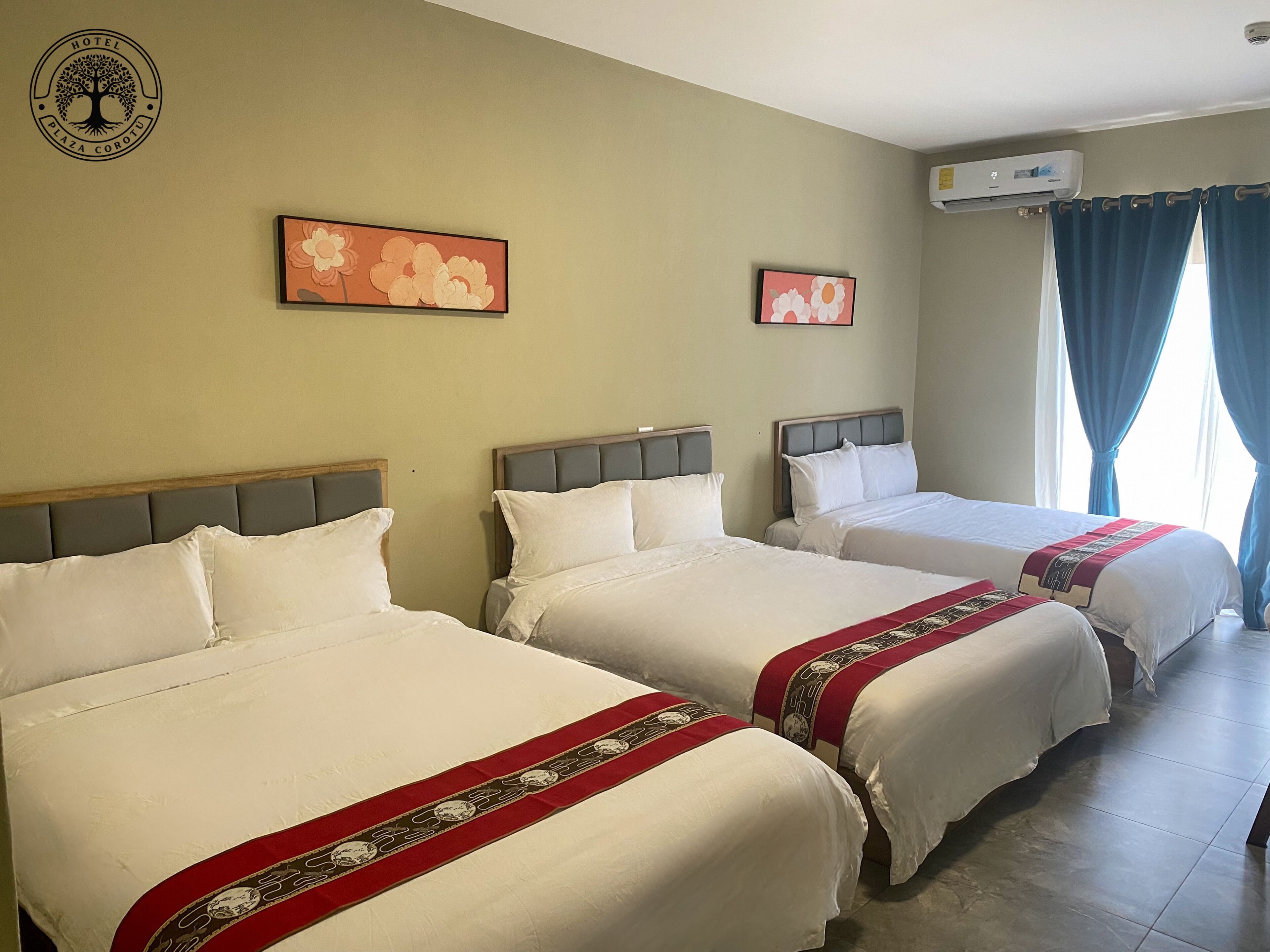 Habitación de hotel con tres camas, aire acondicionado de pared y dos cuadros florales.