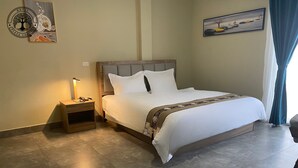 Een keurig opgemaakt bed met hoofdbord, nachtkastje met lamp en een schilderij aan de muur.