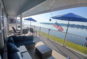 A covered outdoor area with a sofa, chairs, and tables, overlooking a body of water with an American flag. - Beautiful False River 3BR Lake House, 1st & 2nd floor Living rooms & Balconies (New Roads)