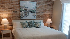 Un dormitorio con pared de ladrillo, un cuadro que muestra una escena de canal, una cama con ropa blanca, dos lámparas de noche y una ventana con persianas.