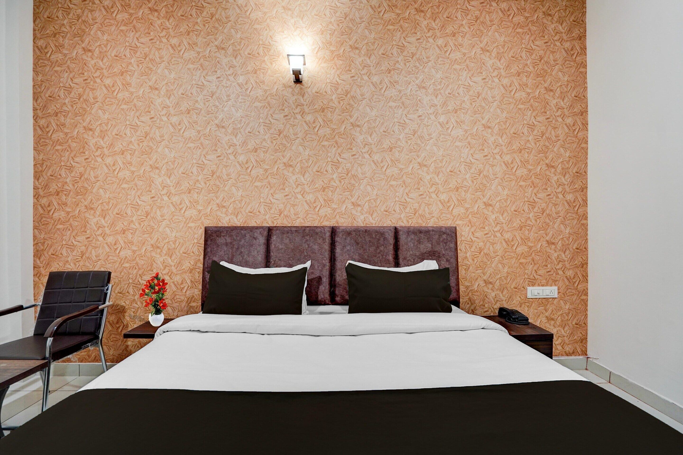 Habitación de hotel con cama, mesita de noche, silla, un florero con flores y una lámpara de pared.