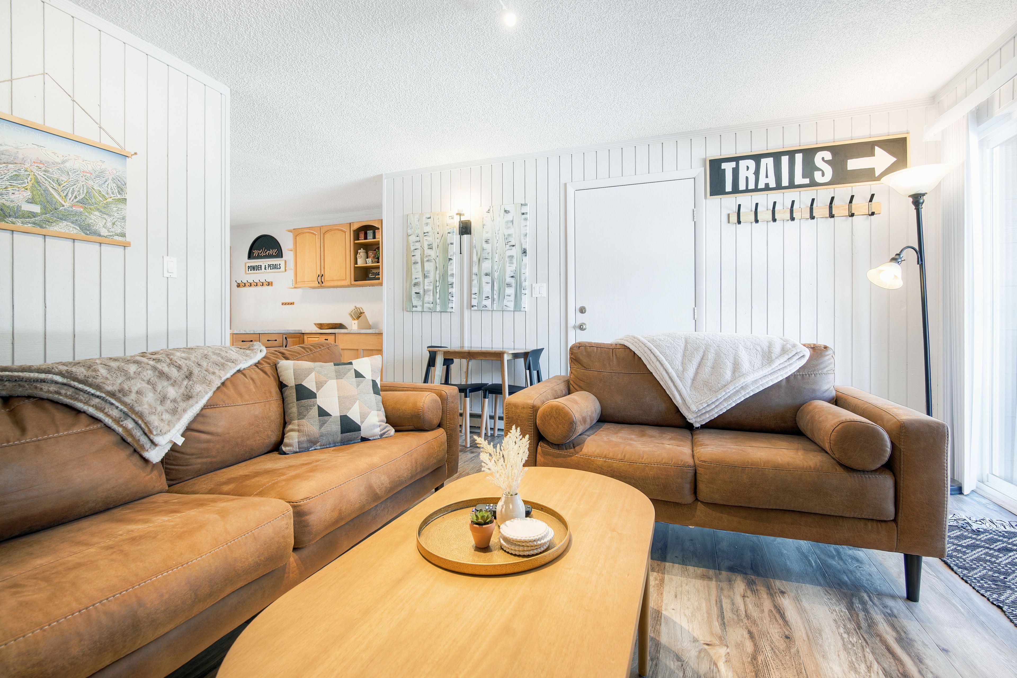 Un salon avec deux canapés en cuir marron, une table basse en bois agrémentée d’objets décoratifs, un panneau « TRAILS » accroché au mur, et un coin repas à l’arrière-plan.
