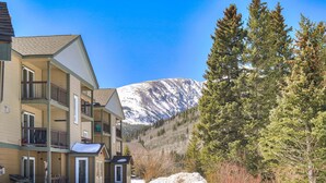 Un hotel de varios pisos con balcones, una montaña nevada al fondo y una zona boscosa.