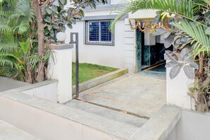 A modern building entrance with a concrete pathway leading to a door, surrounded by lush greenery and a small lawn.