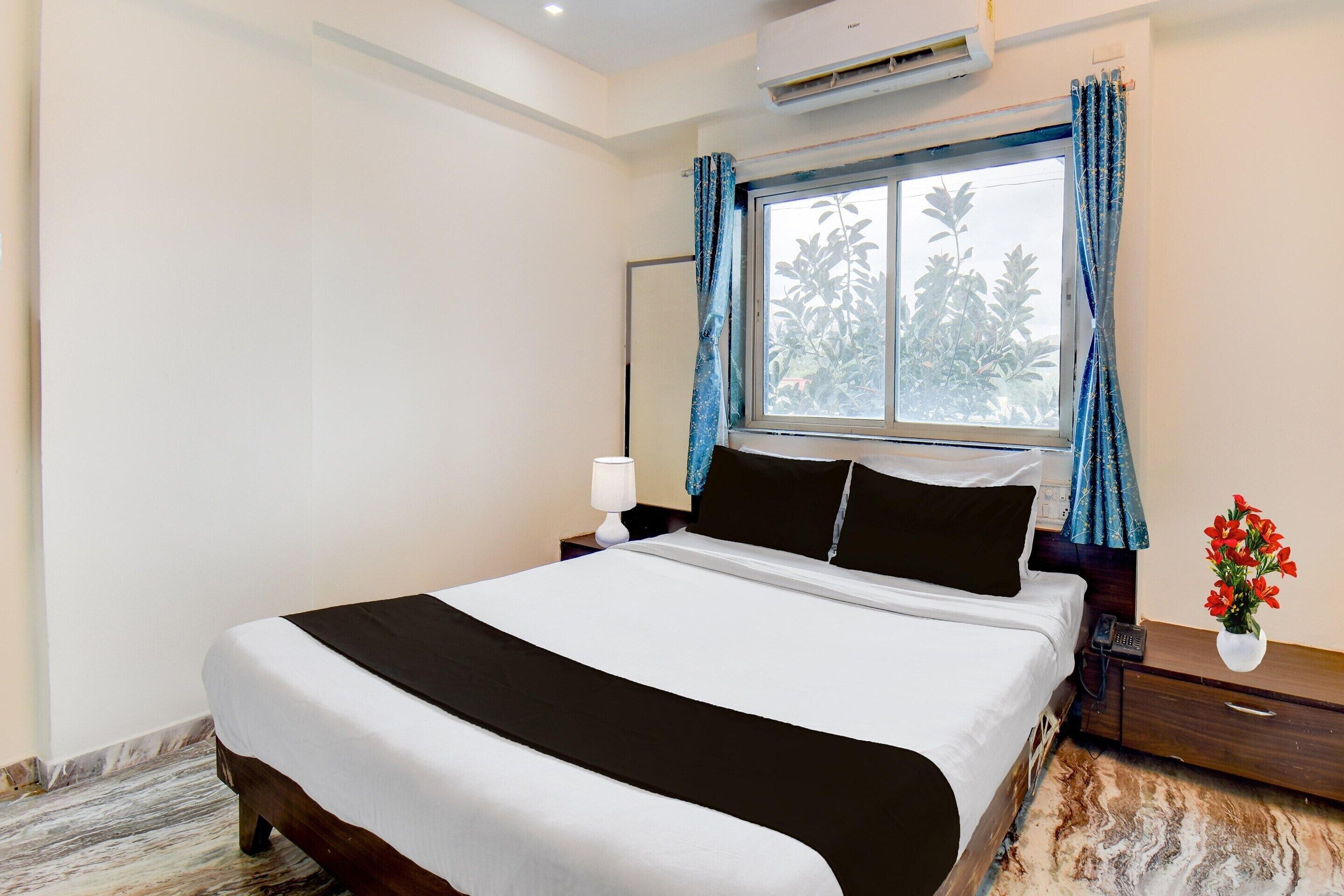 A hotel room with a large bed, a bedside table with a lamp, a nightstand with a phone, a window with curtains, and a vase with flowers.