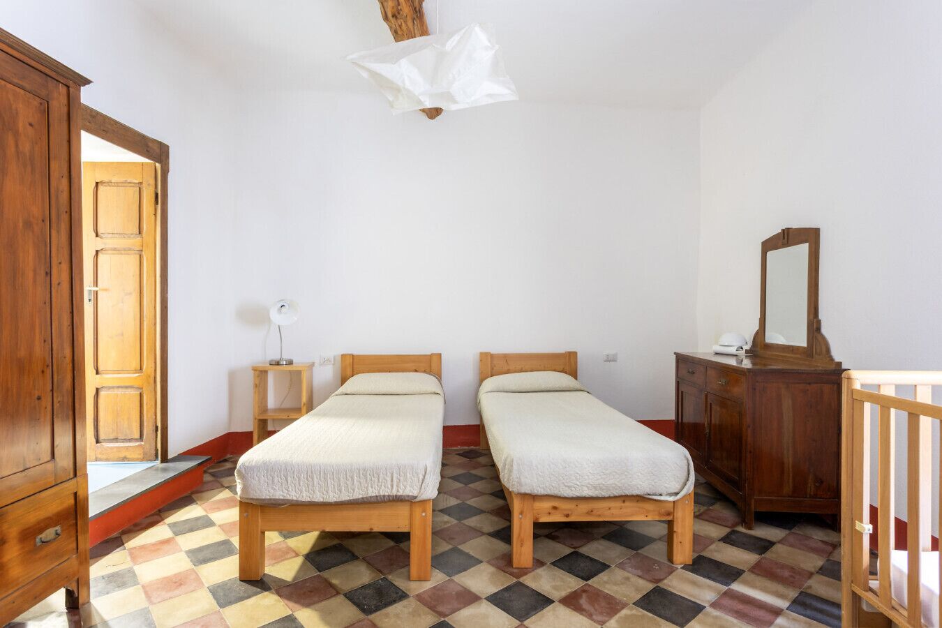 Ein Zimmer mit zwei Einzelbetten, einem Holzschrank, einem kleinen Nachttisch mit Lampe und einem Schachbrettboden.