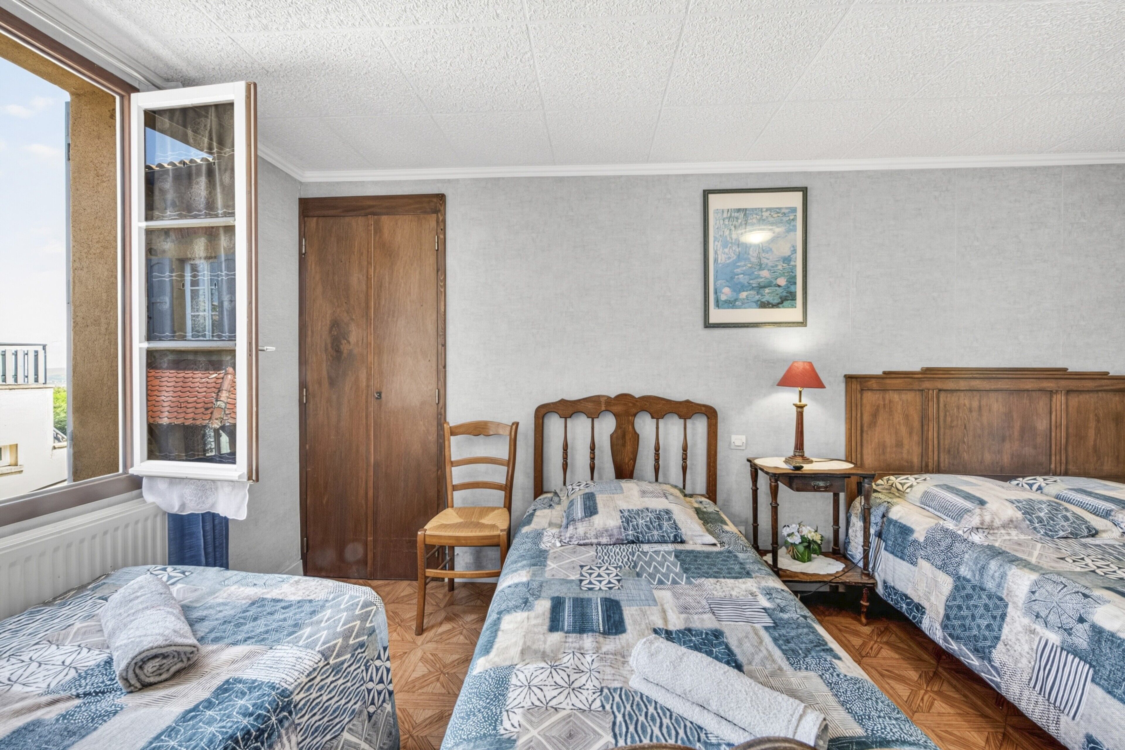 A bedroom with two beds, a wooden wardrobe, a window with a view, a framed picture on the wall, and a small bedside table with a lamp.