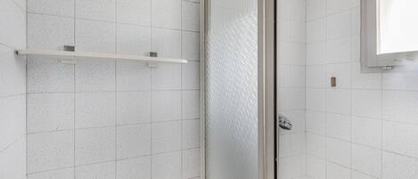 A bathroom with white speckled tiles, a glass shower enclosure, and a small shelf.