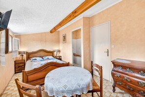 Een slaapkamer met een houten bed, een ronde tafel met een witte tafelkleed, een dressoir met lades en een deur.