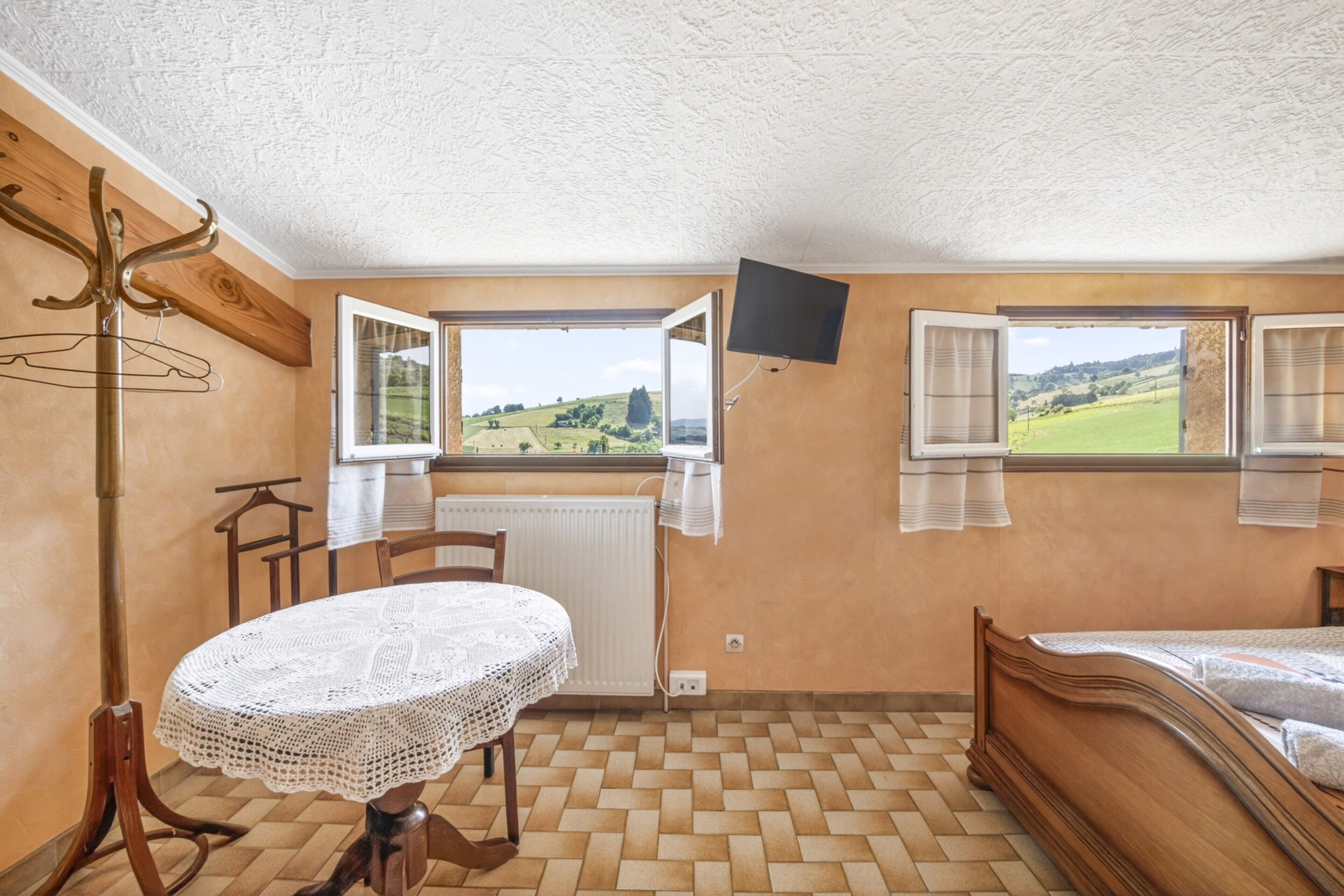 Ein Schlafzimmer mit einem Bett, einem Tisch mit Spitzenbettdecke, einem Kleiderständer, einem Fenster mit Blick ins Grüne und einem Wandfernseher.