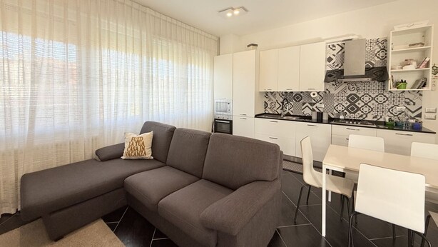 Una sala moderna con un sofá seccional gris, un comedor con una mesa y sillas blancas, y una cocina con salpicadero estampado y muebles de color blanco.