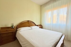 Un letto rifatto con cura, una testiera in legno, un comodino di legno con delle bottiglie e una finestra con tende leggere.