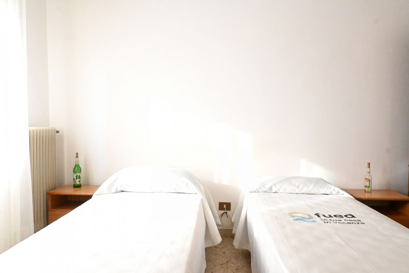 A hotel room with two single beds, a wooden nightstand with a bottle on each, and a radiator on the wall.