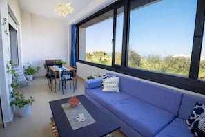 A modern living room with a sofa, a dining table, chairs, and large windows offering a view of greenery.