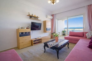 A living room with a TV, wooden furniture, a sofa, and a view of the sea.