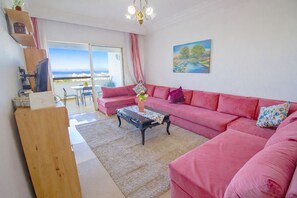 A living room with a pink sofa, a coffee table, a wooden cabinet, and a painting on the wall. - Panoramic Resort Apartment • Sea, Lake & Golf Views • Pools & Tennis (Asilah)