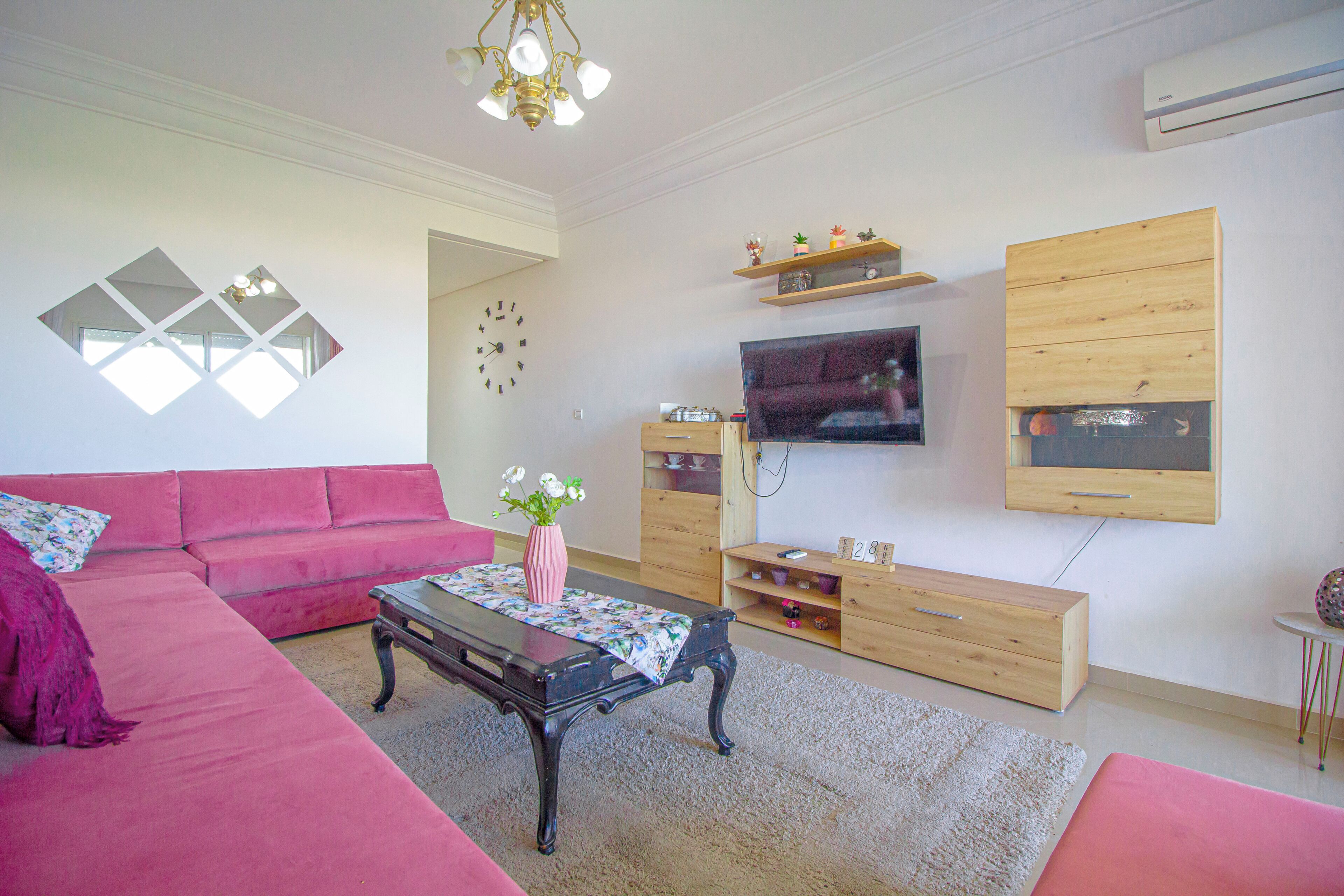 Ein Wohnzimmer mit einem rosafarbenen Sofa, einem Couchtisch, einem Fernseher, Holzböden und einem Wandfernseher.