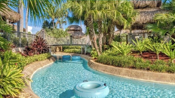 Una piscina con un ponte sospeso, circondata da vegetazione tropicale e costruzioni a tetto di paglia.