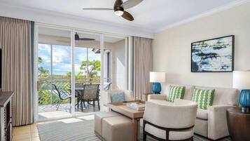 A hotel room with a sofa, armchair, and a dining table. There is a balcony with a table and chairs, and a ceiling fan.
