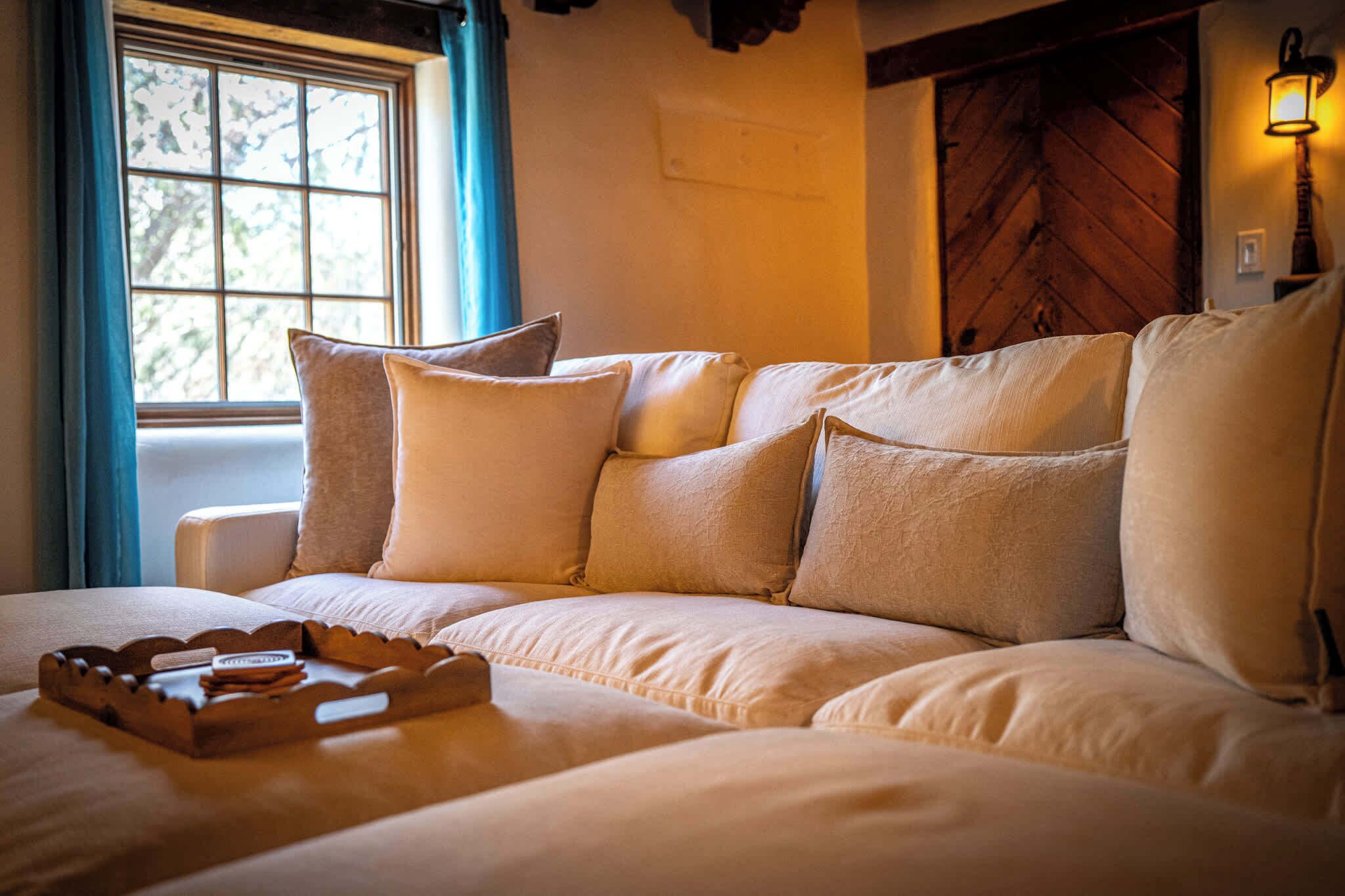 A cozy room with a large sofa, a wooden tray with items on it, a window with blue curtains, and a wall-mounted lamp.