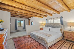 Ein Schlafzimmer mit Holzbalkendecke, einem Bett mit gemustertem Kopfteil, einem Nachttisch mit Lampe und einer Kommode mit Sonnenblumen in einer Vase.