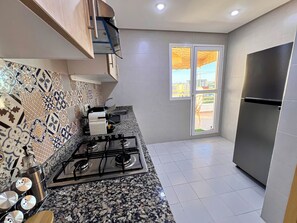A kitchen with a gas stove, a stainless steel refrigerator, and a window with a view of the outside. - Luxury Apartment Sunset – Private Parking (Tanger)