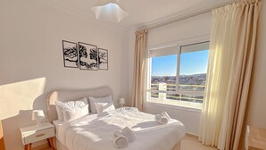 A hotel room with a large bed, a nightstand, a lamp, a chair, and a window with a city view. - Luxury Apartment Sunset – Private Parking (Tanger)
