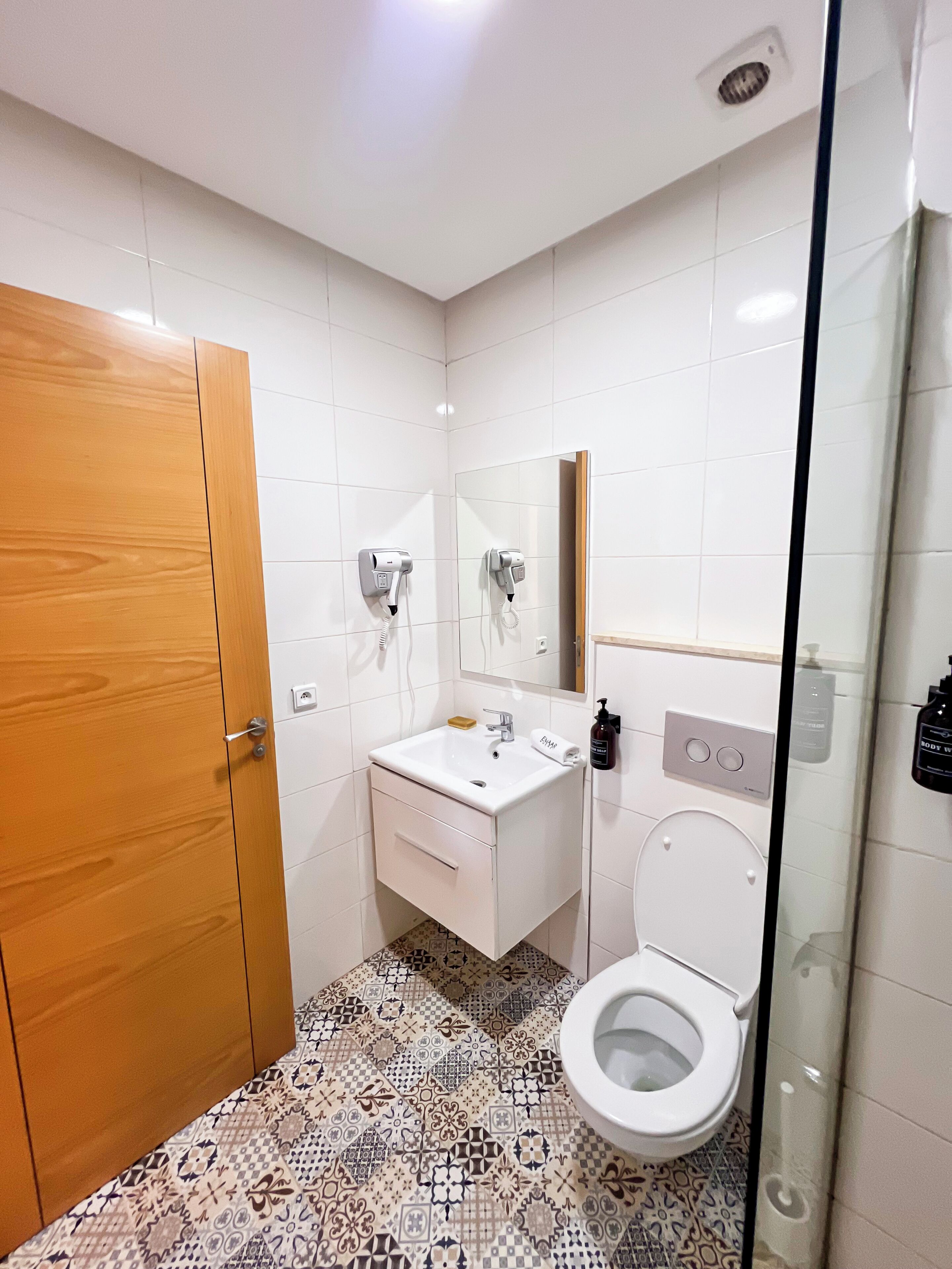 Een badkamer met een houten deur, een witte wastafel met spiegel, een toilet en vloertegels met een patroon.