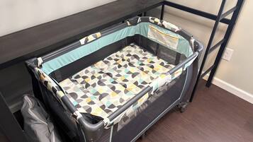 Een draagbaar ledikant met een matras met geometrisch patroon, een gaas zijkant en wielen, geplaatst op een donkere vloer met een muur en een stopcontact op de achtergrond.