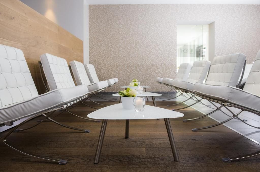 Un espace d’attente moderne avec des chaises blanches rembourrées, une table blanche au centre, ornée d’un vase de fleurs, et un mur à texture.