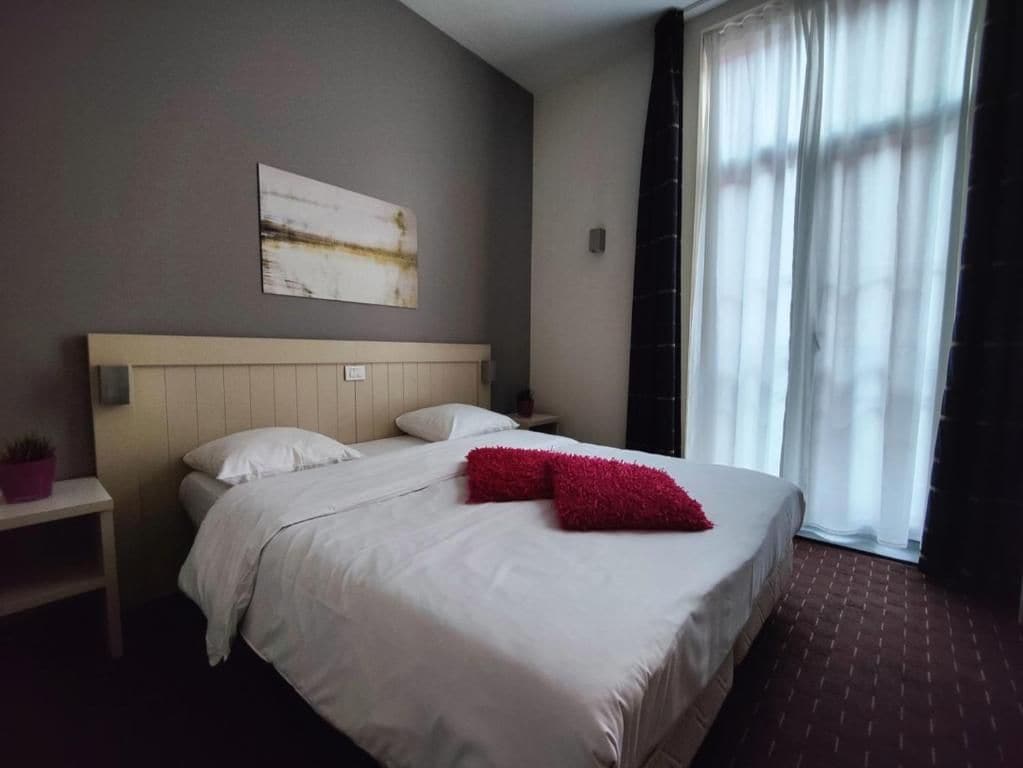 Een hotelkamer met een bed, nachtkastje, een kunstwerk aan de wand en een raam met gordijnen.