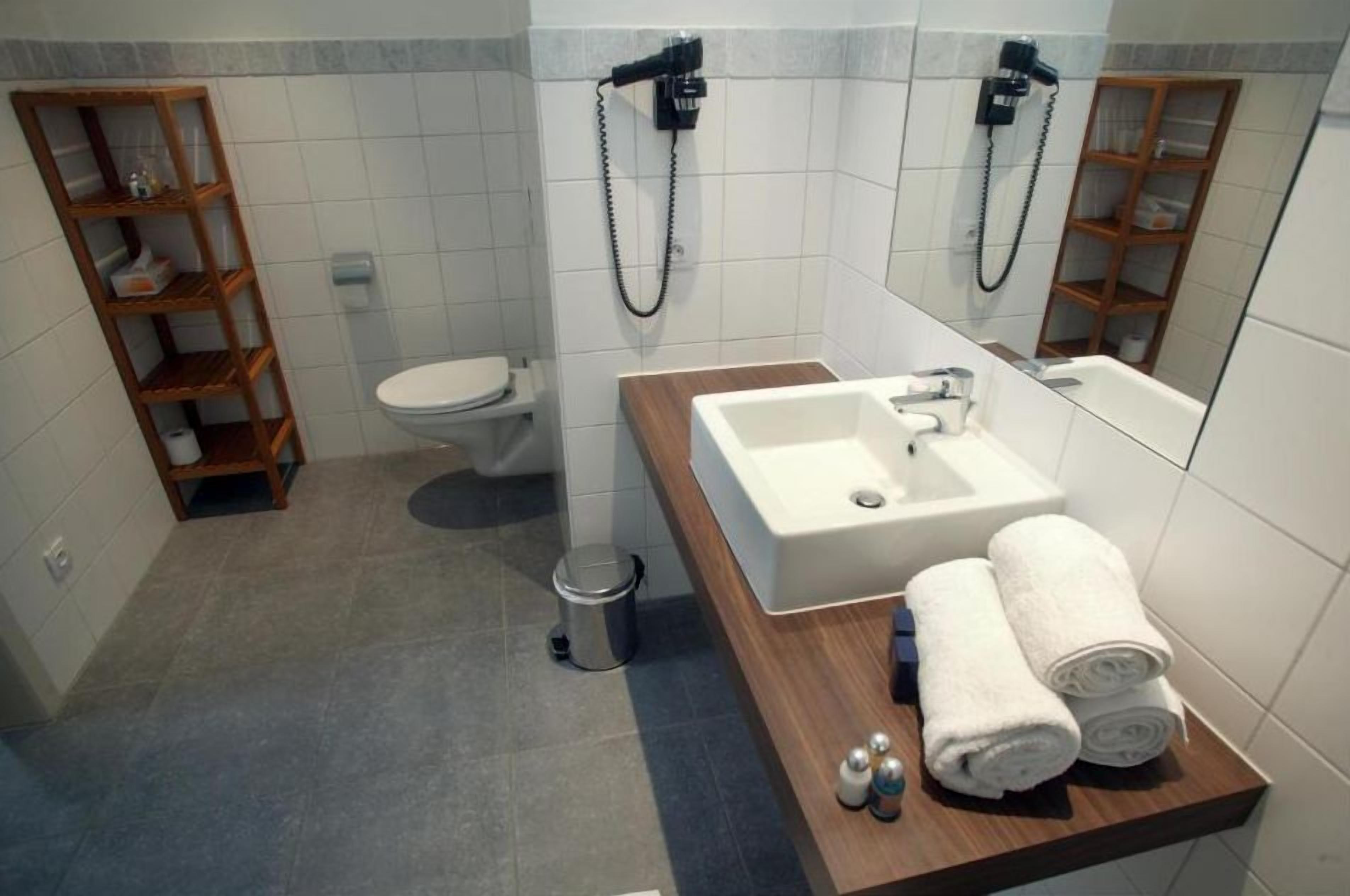 Un baño moderno con inodoro, lavamanos y espejo. Hay estantes de madera y un cubo de basura.