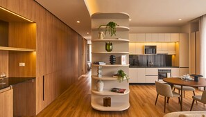 A modern kitchen with wooden cabinetry, a central white shelving unit with decorative items, and a dining area with a round table and chairs. - Kubik Living 8i8 (Bogota)