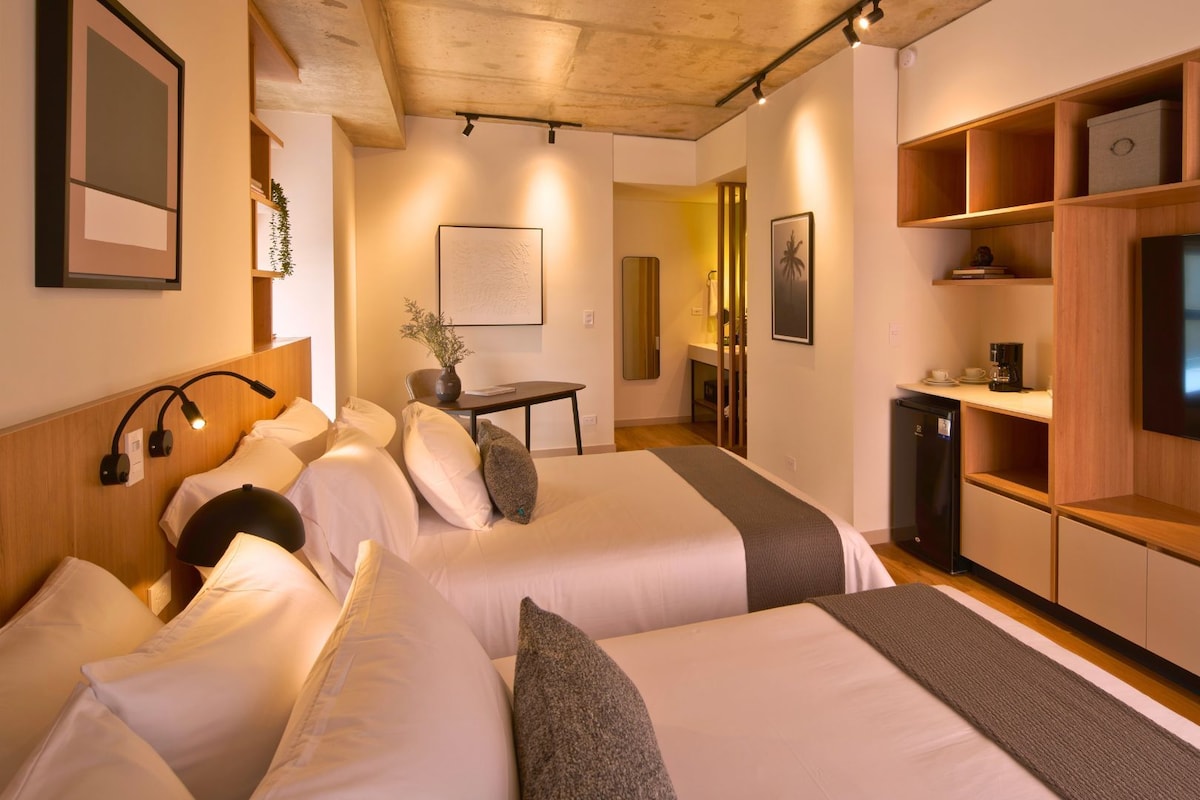 A hotel room with two beds, a desk, a small kitchenette, and a TV.