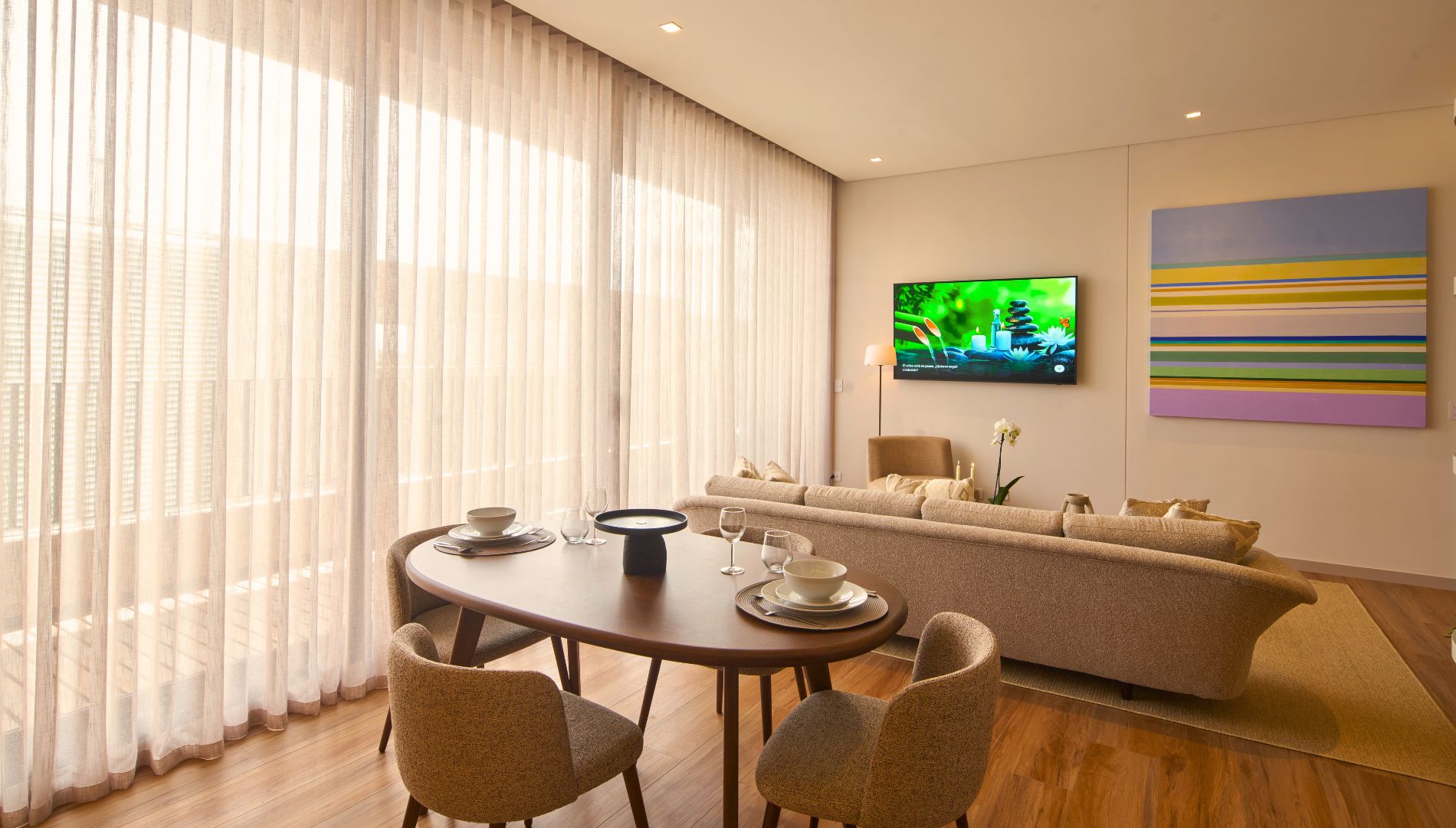 A modern living room with a round dining table, beige sofa, and a flat-screen TV mounted on the wall.