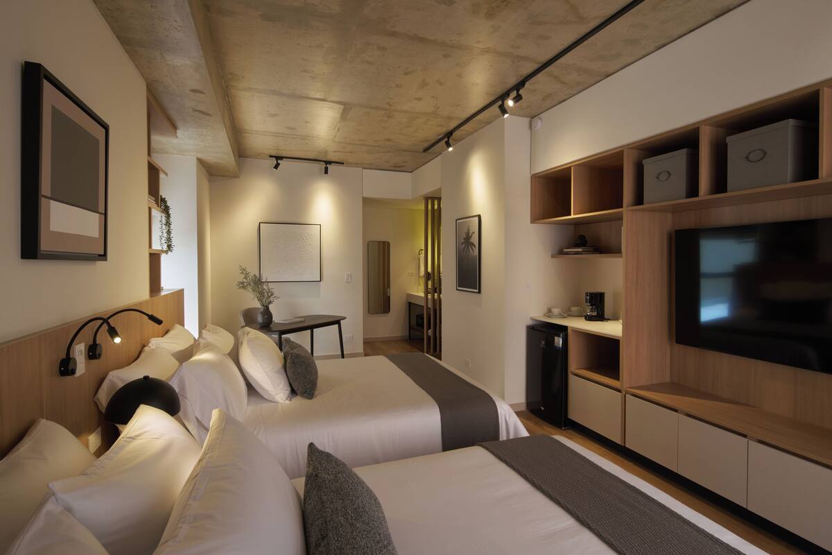 Ett modernt hotellrum med två sängar, en TV, ett litet pentry och ett tak med synliga takbjälkar.