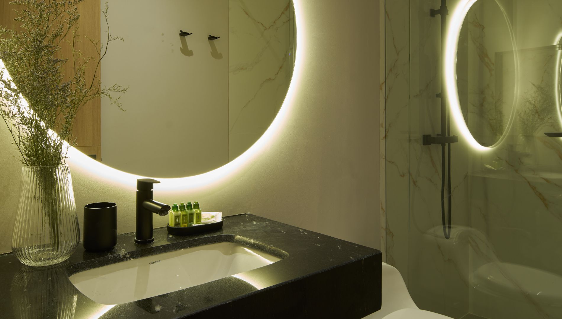 Een badkamer met een zwart wastafel, een spiegel met ronde verlichting, en een vaas met planten.
