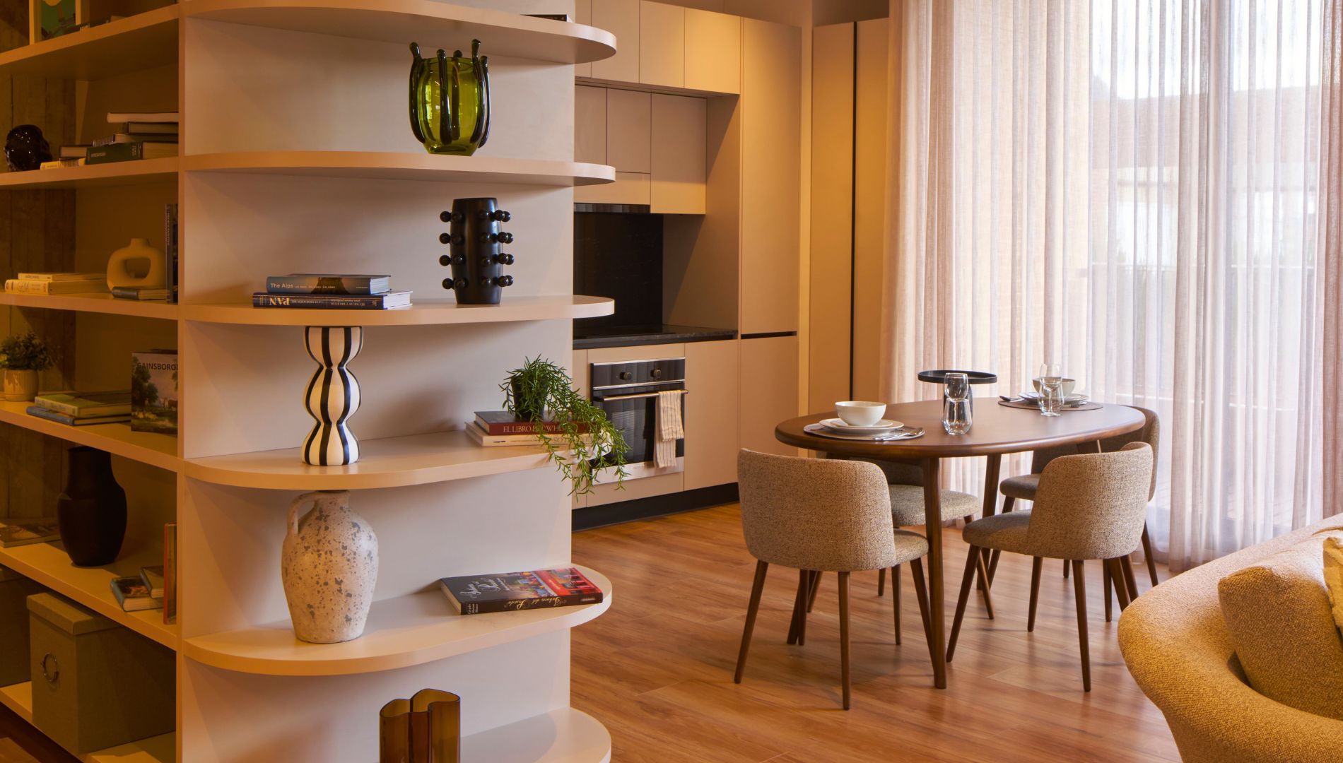 Una sala moderna con una mesa de comedor redonda, varios estantes con libros y objetos decorativos, una zona de cocina con horno y televisión, y un ventanal con cortinas translúcidas.