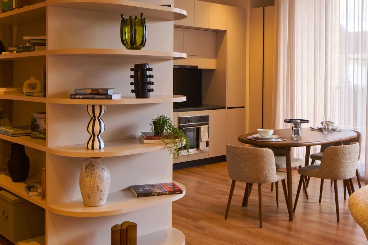 A modern living room with a round dining table, built-in shelving, and a kitchen area with an oven and cabinets.