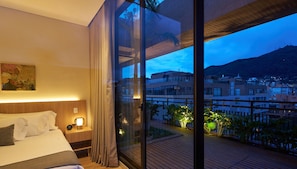 A hotel room with a bed, bedside table, lamp, and a large window with a view of a cityscape and mountains. - Kubik Living 8i8 (Bogota)
