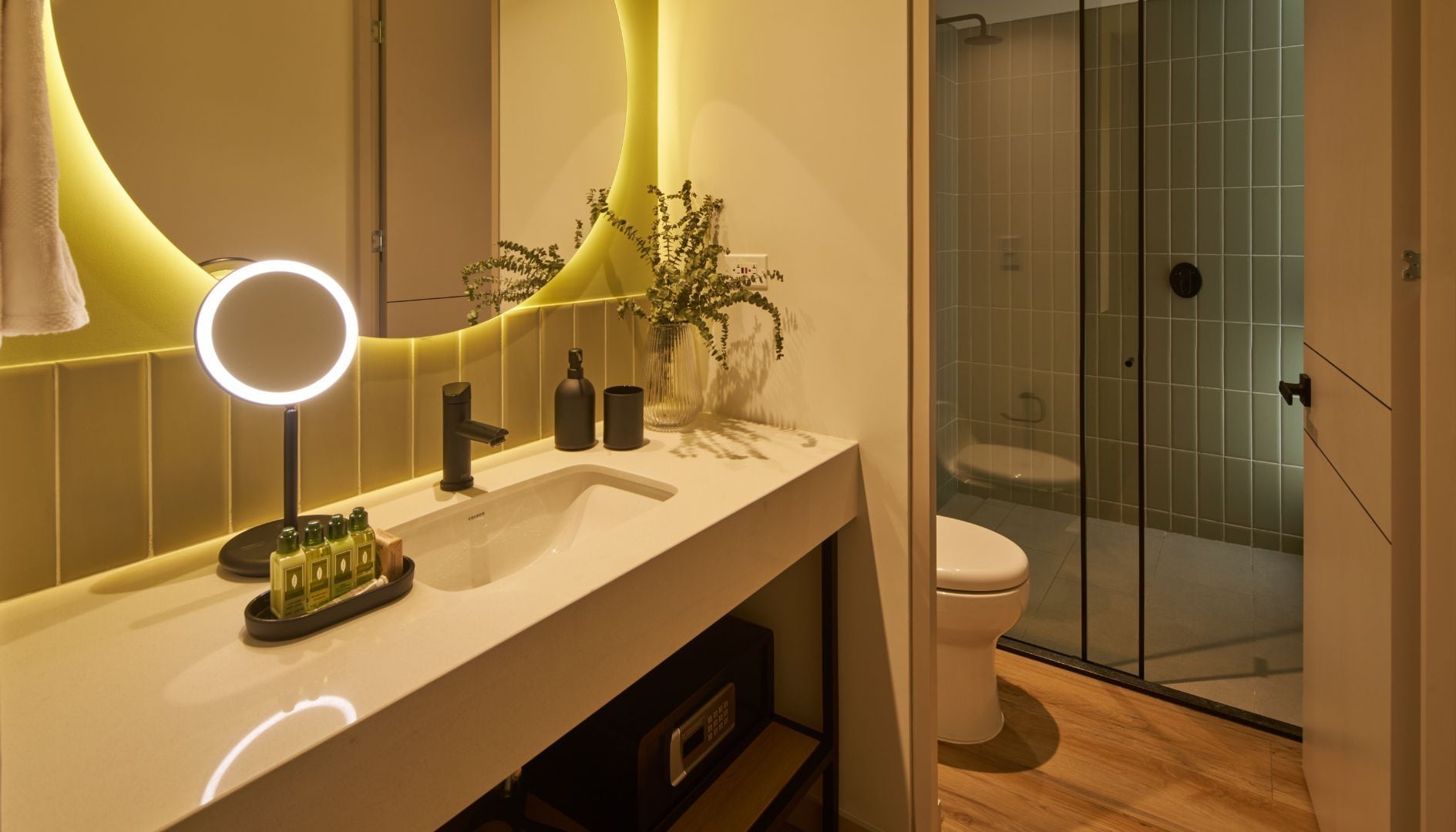 A modern bathroom with a vanity, a round mirror with lights, a toilet, and a glass shower enclosure.