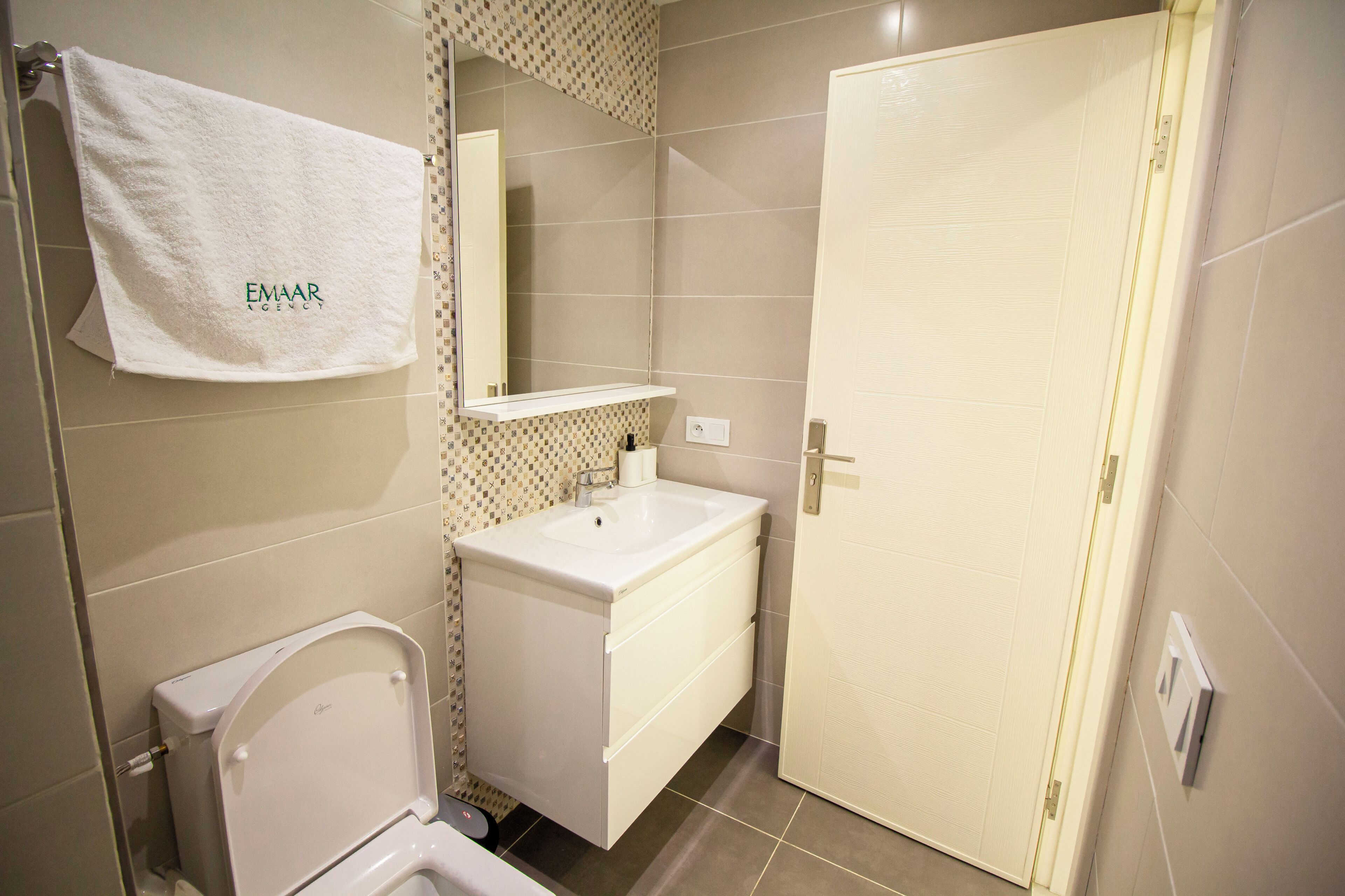 Un baño con inodoro, lavamanos, espejo y perchero para toallas.