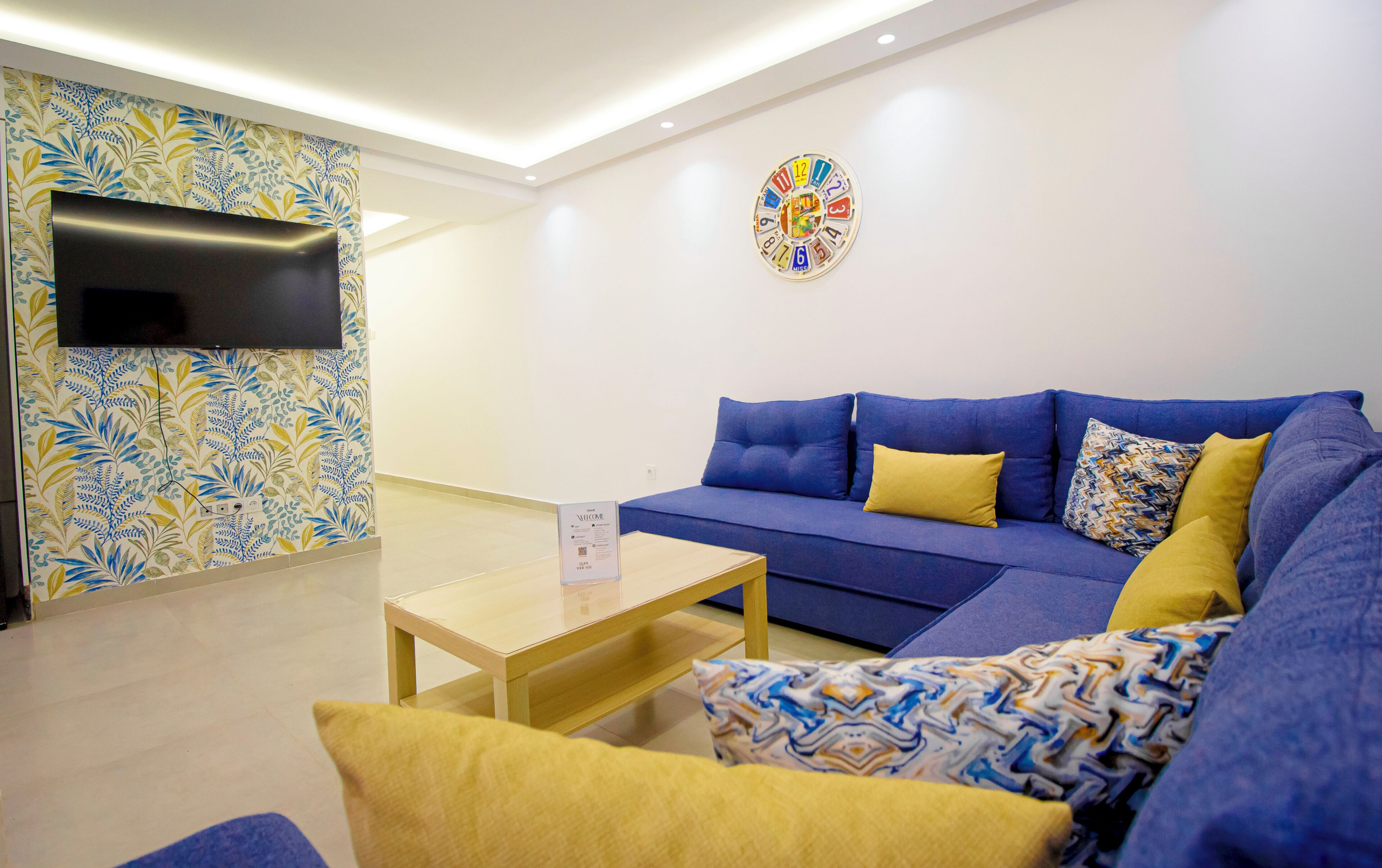 A modern living room with a blue sofa, yellow cushions, a wooden coffee table, a flat-screen TV mounted on a patterned wall, and a colorful clock on the wall.