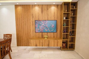 A room with a wooden wall panel, a TV mounted on it, a wooden shelf with decorative items, and a dining table with chairs. - Luxury 2-Bedroom Pool, Quiet & Spacious (Tanger)