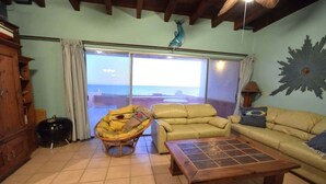 Ein Wohnzimmer mit Meerblick, einem Holztisch, einem gelben Sofa und einem Korbstuhl.