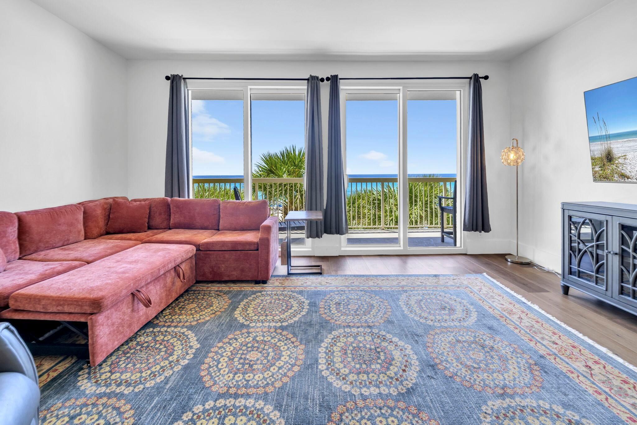 A room with a red sectional sofa, a patterned rug, a balcony with a view of the beach, and a cabinet with a decorative light.