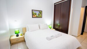 Een net opgemaakt bed met witte lakens, een groene lamp op het nachtkastje, een ingelijst schilderij aan de muur en een houten garderobe.
