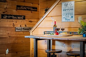 Ein Holzinnenraum mit einem für zwei Personen gedecktem Tisch, einer Weinflasche und Gläsern. An den Wänden befinden sich Schilder mit Sprüchen wie 'Life is what happens between coffee & wine'.