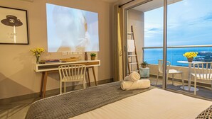 Habitación de hotel con cama, escritorio, silla, laptop, planta y vista al océano.