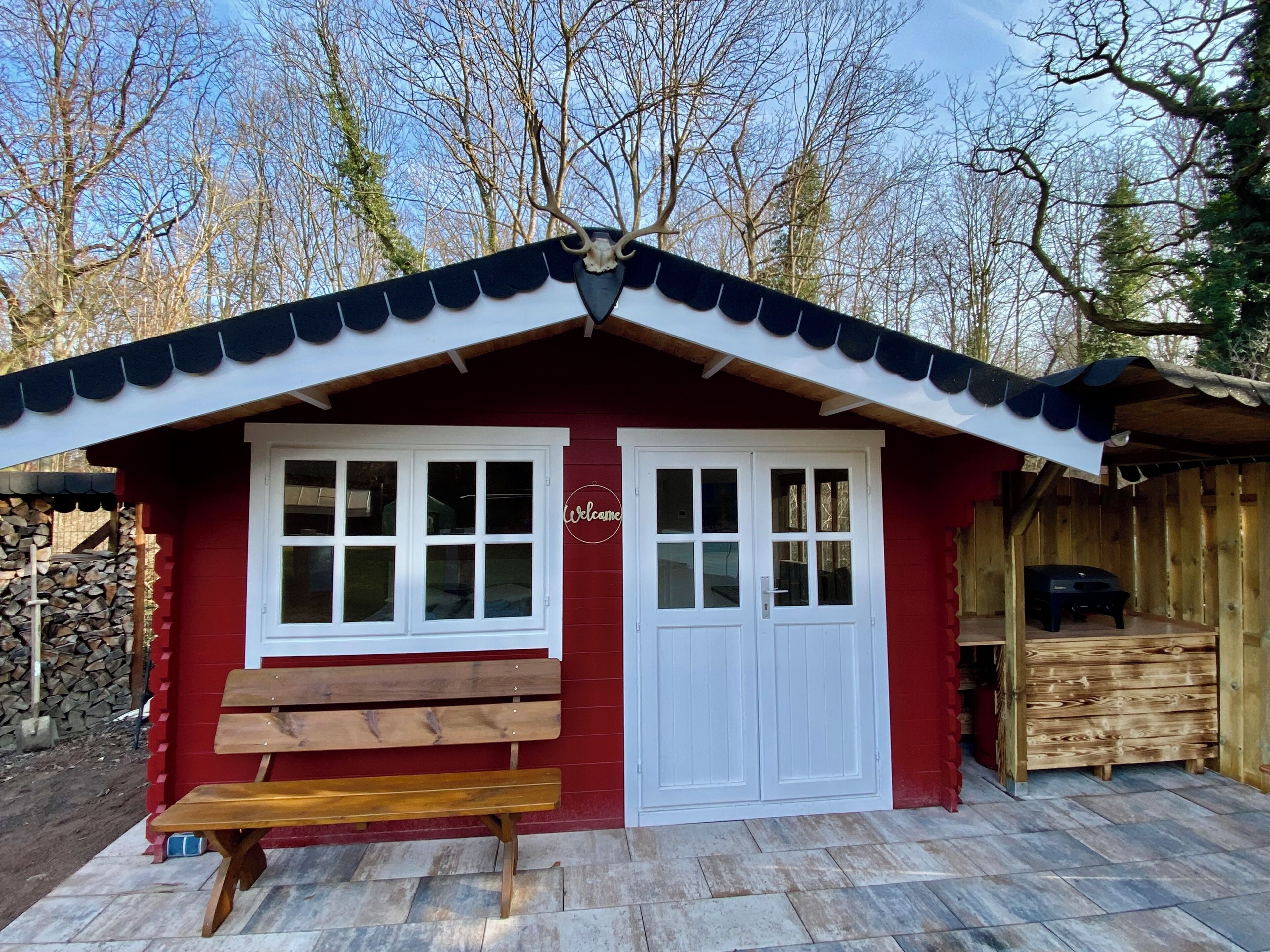 Une cabane en bois rouge avec des fenêtres à cadre blanc, un banc en bois et une pancarte indiquant « Bienvenue ».